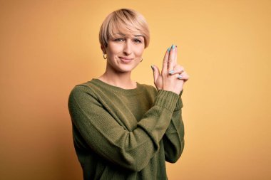 Sarışın, modern kısa saçlı, sarı arka planda günlük süveter giyen, el hareketleriyle sembolik silah tutan, cinayet silahlarıyla oynayan, sinirli yüzlü genç bir kadın.