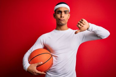 Genç ve yakışıklı Afro-Amerikan sporcusu kırmızı arka planda basketbol topunu tutuyor, olumsuz işaret baş parmakları aşağıda, reddedilme konsepti yok.