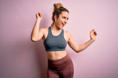 Genç, güzel, sarışın, spor yapan, spor giyinen, pembe arka planda dans eden, mutlu ve neşeli, güler yüzlü, hareketli, rahat ve kendinden emin bir bayan müzik dinliyor.