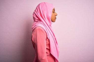 Genç Afrikalı Afro kadın, izole edilmiş pembe arka plan üzerine Müslüman başörtüsü takıyor. Yan tarafa bakıyor, rahat profil pozu veriyor, doğal bir yüzü ve kendine güvenen bir gülümsemesi var..
