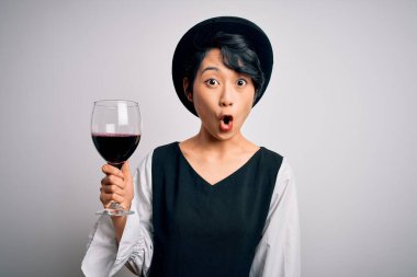 Genç, güzel Asyalı şarap garsonu kız izole edilmiş beyaz arka planda bir bardak kırmızı şarap içiyor. Şaşırmış bir yüzle korkmuş ve korku ifadesinden heyecanlanmış.