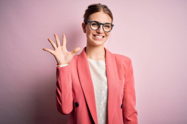 İzole edilmiş pembe arka planda ceket ve gözlük takan genç ve güzel bir kızıl kadın beş numaralı parmaklarıyla işaret ederken kendinden emin ve mutlu bir şekilde gülümsüyor..