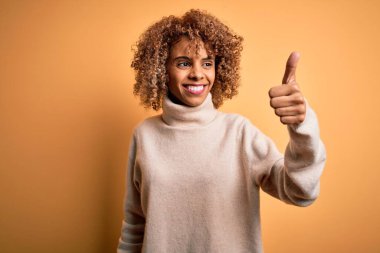 Sarısı arka planda balıkçı yaka kazak giyen güzel Afro-Amerikalı kadın gururlu, gülümseyen baş parmak hareketleriyle kenarda duruyor.