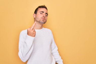 Young handsome man with blue eyes wearing casual sweater standing over yellow background Showing middle finger, impolite and rude fuck off expression