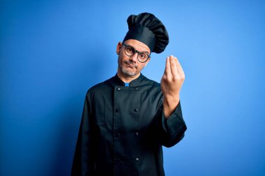 Genç, yakışıklı aşçı üniforması giyen, şapkalı ve soyutlanmış mavi arka planda İtalyan jesti yapan, elleri ve parmakları kendinden emin bir ifade sergileyen bir aşçı.