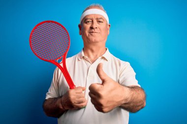 Tenis oynayan orta yaşlı, ağarmış bir sporcu, mavi arka planda raket kullanıyor, gülümsüyor, gülümsüyor, işaret veriyor, parmaklarıyla başparmağını kaldırıyor, mükemmel bir işaret.