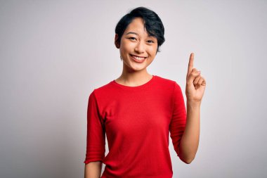 Genç, güzel Asyalı bir kız, günlük kırmızı tişört giyip izole edilmiş beyaz arka planda duruyor. Bir numarayı işaret ediyor ve gülümsüyor. Kendinden emin ve mutlu..