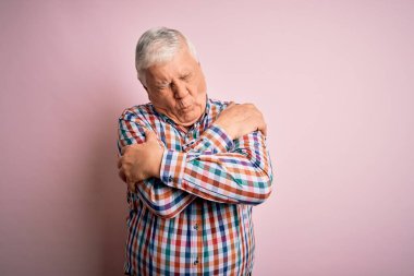 Kıdemli yakışıklı, beyaz gömlekli, pembe arka planda rahat, mutlu ve pozitif, kendinden emin bir şekilde kendine sarılan bir adam. Sevgi ve özgüven.