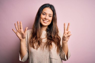 Genç, güzel, esmer kadın pembe arka planda dikilen ve yedinci parmaklarını göstererek gülümseyen, kendinden emin ve mutlu..