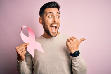 Elinde pembe kanser kurdelesi olan yakışıklı bir adam izole edilmiş arka planda destek sembolü gösteriyor ve gülümseyen bir gülümsemeyle baş parmağıyla işaret ediyor.