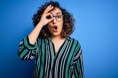 Güzel kıvırcık Arap kadın mavi arka planda çizgili gömlek ve gözlük takıyor. Şaşırmış yüz ifadesiyle şaşırmış, parmaklarından bakan gözler. İnançsız ifade.