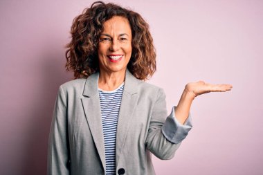 Soyutlanmış pembe arka planda zarif bir ceket giyen, orta yaşlı, güzel bir iş kadını. Gülümseyen, mutlu bir sunum yapan ve avuç içi kameraya bakan..