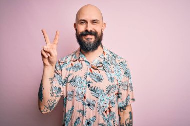 Sakallı ve dövmeli yakışıklı, pembe arka planda sıradan bir çiçek gömleği giyen ve iki numarayı parmaklarıyla gösteren, kendinden emin ve mutlu gülümseyen bir adam..