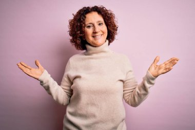 Orta yaşlı güzel kıvırcık saçlı kadın pembe arka planda balıkçı yaka kazak giyiyor. Gülümsüyor, avuçlarını açıyor, sunuyor, reklam yapıyor, karşılaştırıyor ve dengede duruyor.