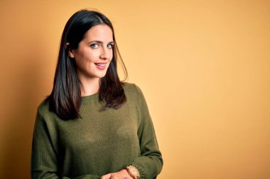 Sarı arka planda yeşil süveter giyen, mavi gözlü genç esmer kadın ellerini birleştirip gülümseyerek gevşemiş ve neşeliydi. Başarı ve iyimser