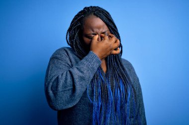 Mavi arka planda günlük süveter giyen Afrikalı Amerikalı artı cüsseli kadın yorgun burnu ve gözlerini ovuşturup yorgunluk ve baş ağrısı çekiyor. Stres ve hüsran kavramı.