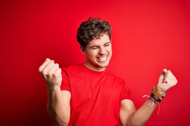 Sarışın, kıvırcık saçlı, kırmızı arka plan üzerine gündelik tişört giyen çok mutlu ve heyecanlı genç bir adam kolları havada, gülümseyen ve başarı için çığlık atan, kazanan bir jest yapıyor. Kutlama konsepti.