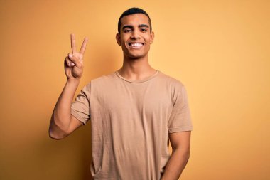 Sarı arka planda dikilmiş gülümseyen ve parmaklarını zafer işareti yaparken gösteren gündelik tişörtler giyen genç, Afro-Amerikan bir adam. İki numara..