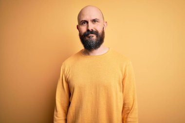 Sarı arka planda dikilen sıradan bir kazak giyen sakallı yakışıklı kel adam yüzünde ciddi bir ifadeyle rahatlamış. Basit ve doğal bir şekilde kameraya bakıyorsun..