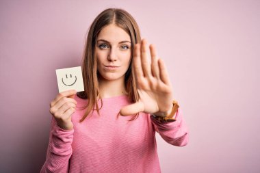 Genç, güzel, kızıl saçlı bir kadın elinde gülümseyen emoji mesajıyla elinde hatırlatıcı kağıtla elinde dur işareti ile ciddi ve kendinden emin bir ifadeyle savunma jesti yapıyor.