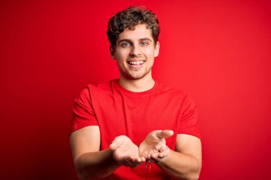 Sarışın, kıvırcık saçlı, kırmızı arka planda sıradan tişörtler giyen, elleri avuçlarında gülümseyen, el hareketi yapan genç bir adam. Tutun ve koruyun