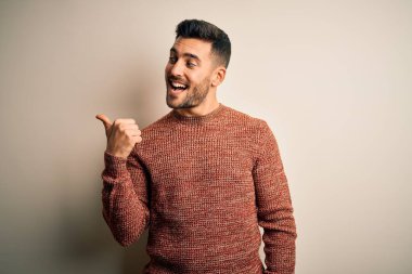 İzole edilmiş beyaz arka planda dikilen, gülen yüzlü ve baş parmağıyla yüzünü gösteren, sıradan bir süveter giyen genç yakışıklı adam..