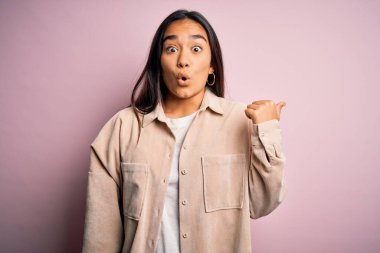 Genç ve güzel Asyalı kadın pembe arka planda dikilen rahat bir gömlek giyiyor. El parmağı yana dönük, ağzı açık, şaşkın bir ifade..