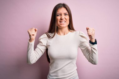 Mavi gözlü genç bir kadın pembe arka plan üzerine gündelik beyaz tişört giyiyor kızgın ve kızgın yumruklar sallarken öfkeyle bağırıyor. Öfke ve agresif kavram.