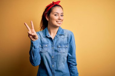 Genç, güzel, esmer, günlük kot gömlek ve saç mendili giyen bir kadın. İki numaralı parmaklarıyla kendini beğenmiş ve mutlu bir şekilde iki numarayı işaret ediyor..