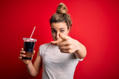 Güzel sarışın kadın kırmızı zemin üzerinde ferahlatıcı kola içerek kamerayı işaret ediyor ve sen, el işareti, ön taraftan pozitif ve kendinden emin bir jest.