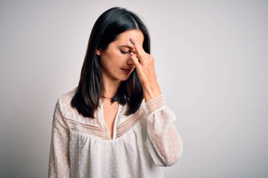 Mavi gözlü genç esmer kadın izole edilmiş beyaz arka planda günlük tişört giyiyor. Burnu ve gözlerini ovuşturup yorgunluk ve baş ağrısı çekiyor. Stres ve hüsran kavramı.