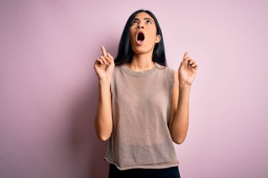 Pembe arka planda süveter giyen genç ve güzel İspanyol moda kadını şaşırmış ve şaşırmış bir şekilde yukarı bakıyor ve parmaklarıyla işaret ediyor ve kollarını kaldırıyor..