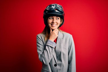 Kırmızı arka planda motosiklet kaskı takan genç, sarışın, güzel bir motosikletçi kadın. Elleri çenesinde, soru sormayı düşünüyor. Düşünceli bir ifade. Düşünceli bir yüzle gülümsüyor. Şüphe kavramı.