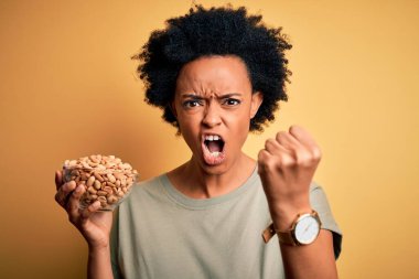 Genç Afrikalı Afro kadın, kıvırcık saçlı, elinde tuzlu fıstıkla sinirli sinirli bağırıyor, çıldırıyor ve el kaldırarak bağırıyor.