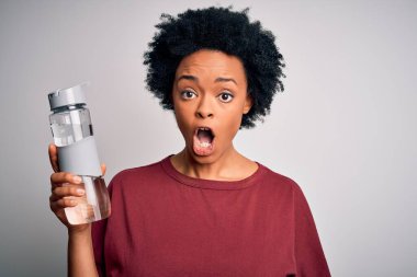 Kıvırcık saçlı genç Afrikalı Afro kadın ferahlamak için bir şişe su içiyor. Şaşırmış bir yüz ifadesinden korkuyor ve heyecanlanıyor.
