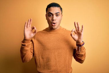 İzole edilmiş sarı arka planda dikilen sıradan süveter giyen genç Brezilyalı adam şaşırmış ve şaşırmış görünüyor. Tamam sembolünü parmaklarıyla yapıyor. Çılgın ifade