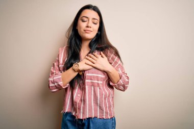 İzole edilmiş arka plan üzerinde sıradan çizgili tişört giyen genç esmer kadın elleri göğsünde, gözleri kapalı ve yüzünde minnet dolu bir jest ile gülümsüyor. Sağlık kavramı.