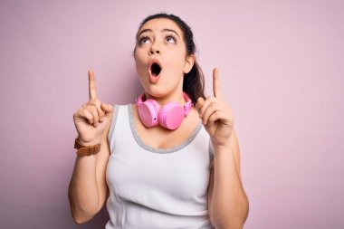 Pembe izole arka planda spor kulaklığı takan esmer, genç bir kadın şaşırmış ve şaşırmış bir şekilde parmaklarıyla işaret ediyor ve kollarını kaldırıyordu..