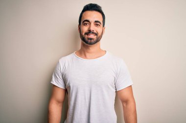 Sakallı, sıradan tişörtlü, beyaz arka planda mutlu ve havalı bir gülümsemesi olan genç yakışıklı bir adam. Şanslı kişi..