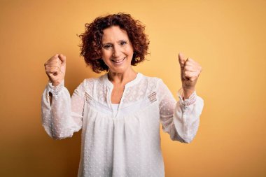 Orta yaşlı güzel kıvırcık saçlı kadın sarı arka plan üzerine gündelik bir elbise giyip kollarını kaldırarak ve açık gözlerle başarıyı kutluyor. Kazanan konsept.