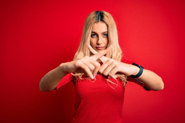 Genç, güzel, sarışın bir kadın, günlük tişört giyiyor, kırmızı arka plan üzerinde duruyor, reddedilişin olumsuz işaret vermesi için parmaklarını çapraz yapıyor.