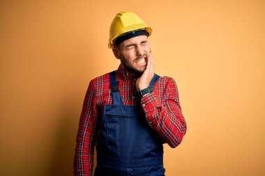 İnşaat üniforması giyen ve sarı yalıtılmış arka plan üzerine güvenlik kaskı takan genç bir müteahhit diş ağrısı ya da diş ağrısı yüzünden ağız ve acı dolu bir ifadeyle birbirine dokunuyor. Dişçi