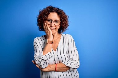 Orta yaşlı, kıvırcık saçlı, rahat çizgili tişörtlü, izole bir arka plan giyen güzel bir kadın kollarını kavuşturup depresyondan sıkılmış ve yorgun göründüğünü düşünüyor..