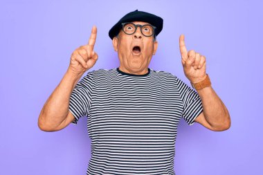 Kıdemli yakışıklı, beyaz bereli ve gözlüklü adam mor arka plan üzerinde şaşkın şaşkın ve şaşırmış bir şekilde yukarı bakıyor ve parmaklarıyla işaret ediyor ve kollarını kaldırıyor..