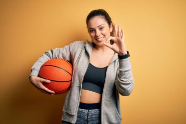 Sarı arka planda basketbol topu tutan güzel esmer bayan. Tamam işareti yapıyor. Parmaklarıyla, mükemmel bir sembol.