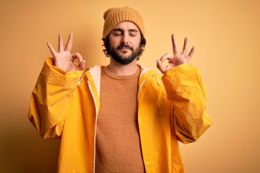 Sarı arka planda yağmurluk giyen sakallı yakışıklı genç adam rahatla ve gözün kapalı gülümse parmaklarınla meditasyon hareketi yap. Yoga konsepti.