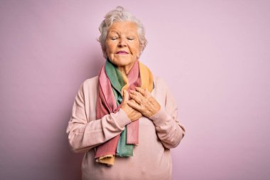 Son sınıf güzeli gri saçlı kadın pembe arka planda günlük süveter ve eşarp giyiyor elleri göğsünde, gözleri kapalı ve yüzünde minnet dolu bir jest ile gülümsüyor. Sağlık kavramı.