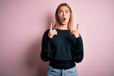 Pembe izole edilmiş arka planda güzel sarışın kadın şaşırmış ve şaşırmış bir şekilde yukarı bakıp parmaklarıyla işaret ediyor ve kollarını kaldırıyordu..