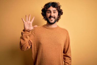 Sakallı, sıradan bir süveter giyen, sarı arka planda duran, beş numaralı parmaklarıyla işaret eden, kendinden emin ve mutlu gülümseyen genç yakışıklı bir adam..