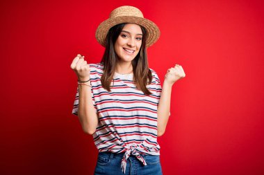 Kırmızı arka plan üzerinde çizgili tişört ve yaz şapkası giyen genç ve güzel esmer kadın kollarını kaldırarak ve açık gözlerle başarıyı kutluyor ve şaşırıyor. Kazanan konsept.